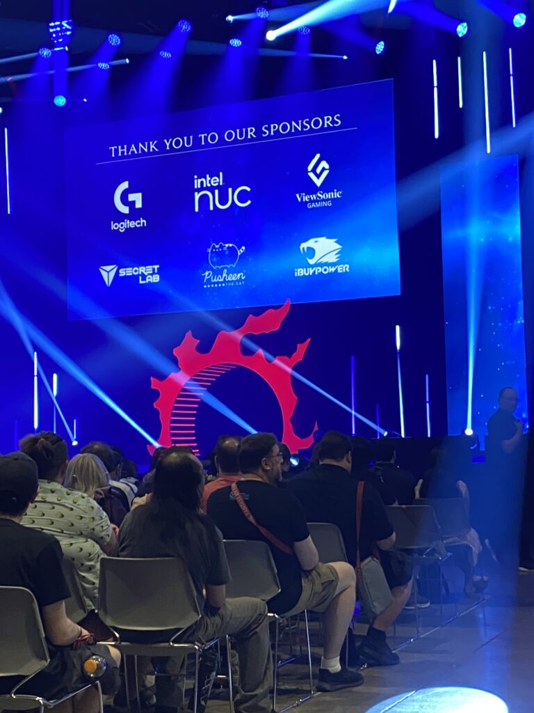 Picture of the keynote stage at Final Fantasy XIV Fan Fest in Las Vegas. A red meteor-logo arch is on the stage, with a large screen currently listing event sponsors above it.