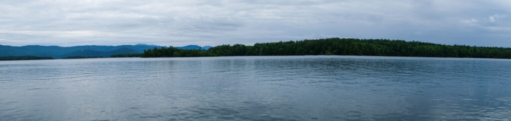 A panorama of Squam Lake. The sky is cloudy and the water is calm.